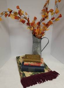 lot 114 image: Fringed Floral Tapestry Table Runner 13in W x 62in L, Rustic Embossed Metal Pitcher with Fall Folliage Arrangement, and 3 Vintage Books
