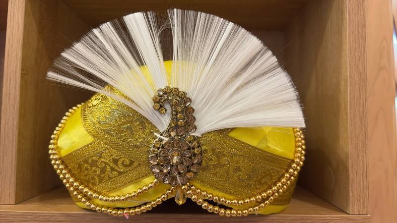lot 1905 image: This is a traditional yellow and gold wedding turban, also known as a safa, pagri, or thalapa, adorned with a decorative jewel brooch and a white feather.