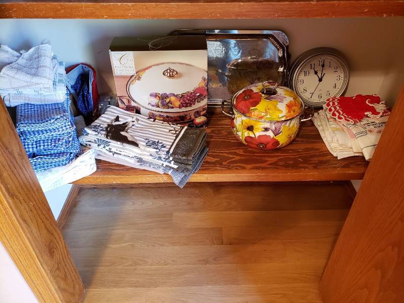lot 3312 image: Lot of Kitchen Items on Bottom Shelf of Food Pantry - Linens, Decorative Servers, Clock, and Pot Holders